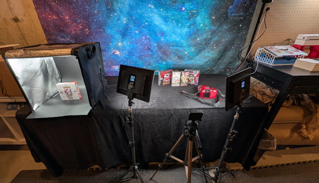 Holo Reserve product photography studio setup in basement — lightbox with Pokemon 151 box, two LED panels on tripods, DSLR camera, and galaxy space backdrop