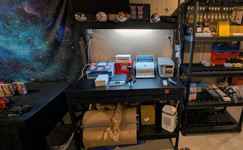 Holo Reserve shipping and packing station — WEN workbench with thermal label printer, postage scale, water-activated tape dispenser, and Pokeball display shelf above